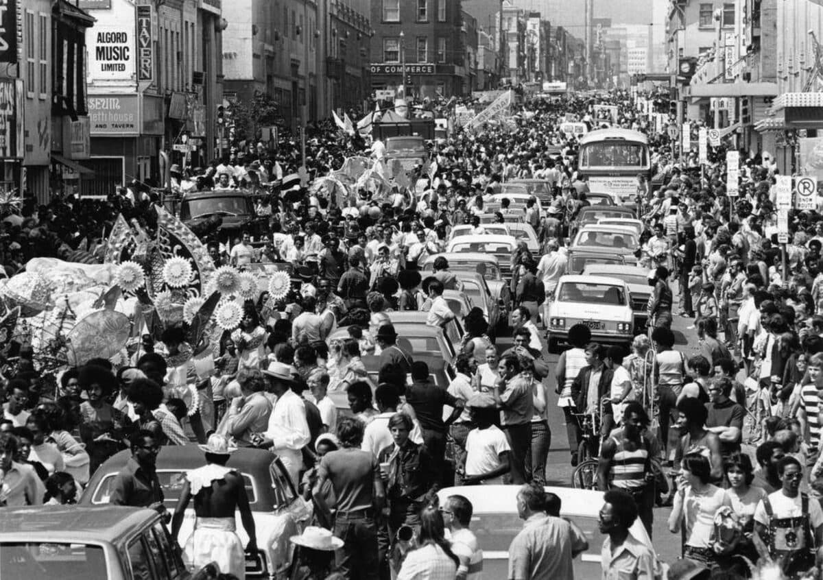Black-and-white photograph of a street packed with people and cars during a festival, with costumed revellers on the left