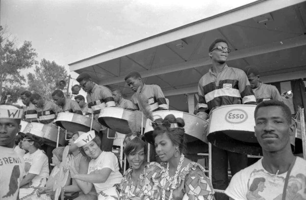 Black-and-white photograph of a steelband in matching jackets playing steel pans marked Esso, with onlookers in the foreground