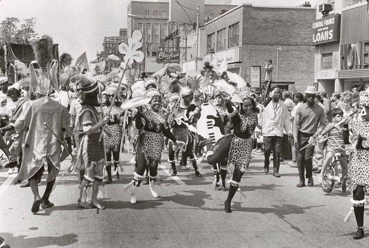 Black-and-white photograph of a parade on a city street, masqueraders in leopard-print costumes with feathered headdresses, crowds on both sides