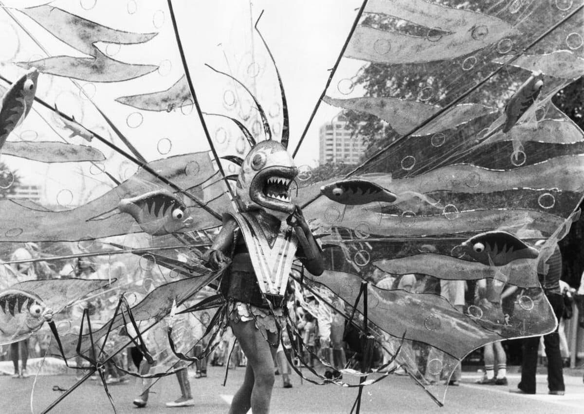 Black-and-white photograph of a masquerader in a fish-headed costume with huge transparent wings patterned with fish and bubbles