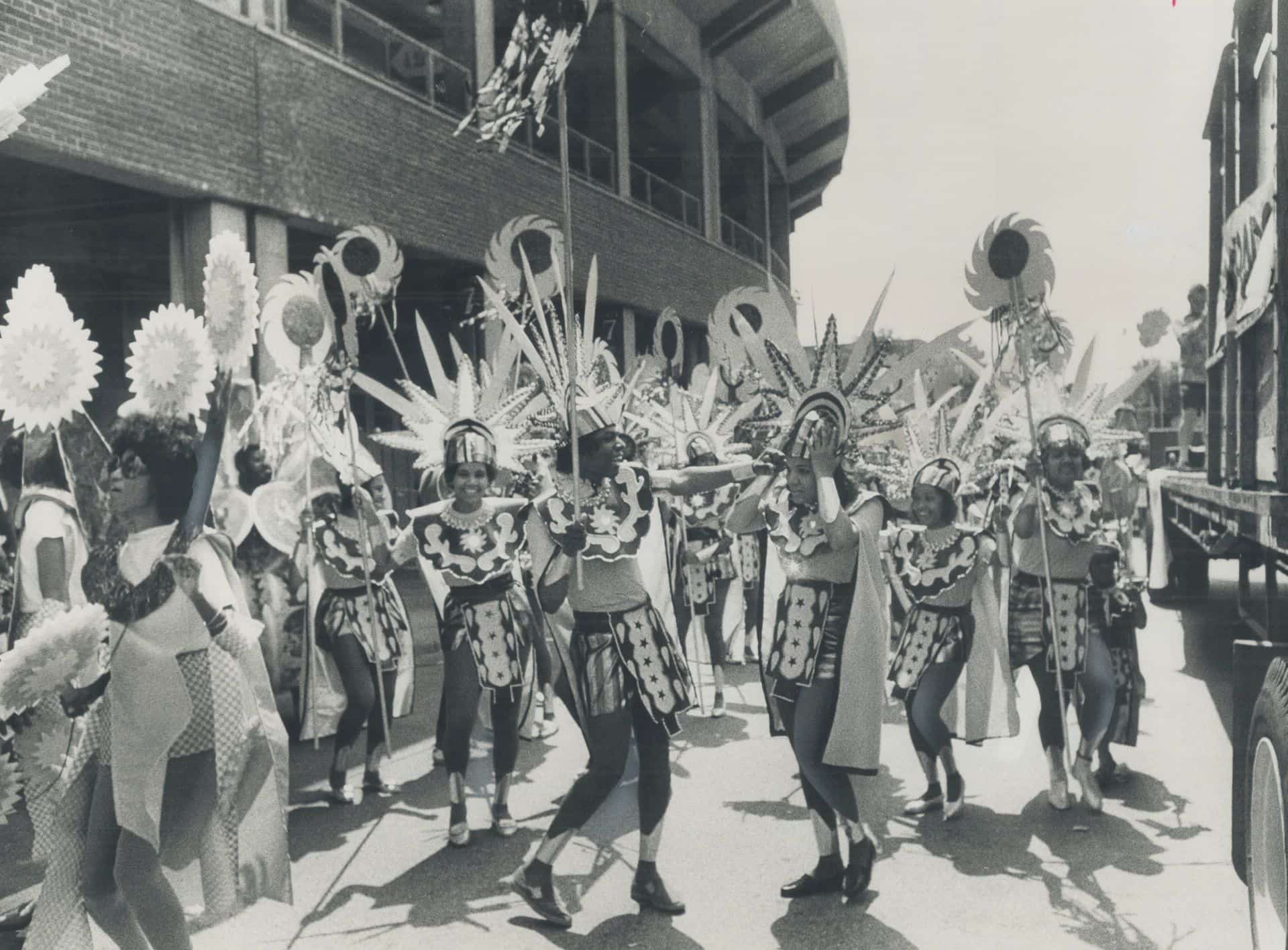 Black-and-white photograph of masqueraders in sun-shaped headdresses and patterned costumes dancing along a street beside a stadium